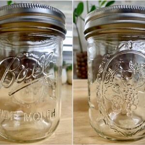 May include: Two clear glass Ball wide mouth jars with silver lids. One jar has the Ball logo and the words "Wide Mouth" printed on the side. The other jar has a grapevine design.