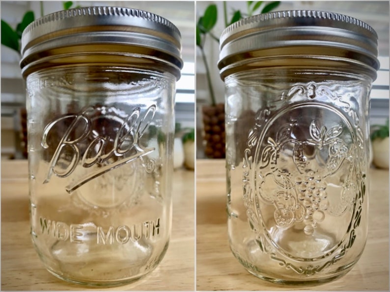 May include: Two clear glass Ball wide mouth jars with silver lids. One jar has the Ball logo and the words "Wide Mouth" printed on the side. The other jar has a grapevine design.