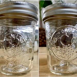 May include: Clear glass Ball jar with a silver metal lid. The jar has embossed fruit and leaf designs, with the word "Ball" in cursive. The jar is marked with measurements in ounces and is made in the USA.