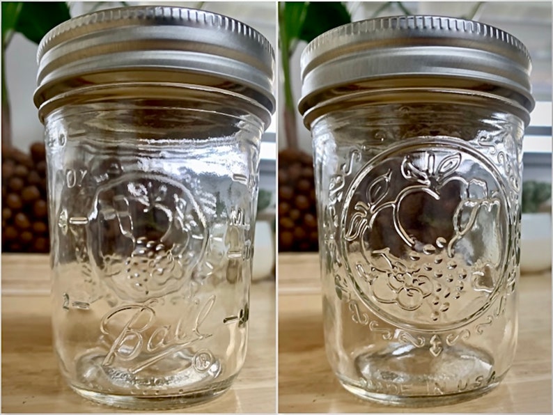 May include: Clear glass Ball jar with a silver metal lid. The jar has embossed fruit and leaf designs, with the word "Ball" in cursive. The jar is marked with measurements in ounces and is made in the USA.