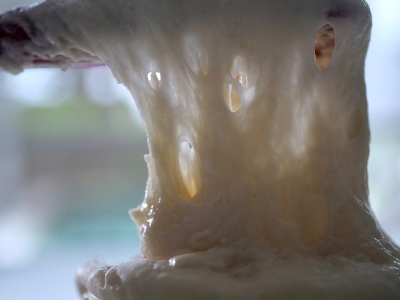 May include: Close-up of white dough being stretched, showing its elasticity and texture. The dough is soft and airy, with small holes throughout.