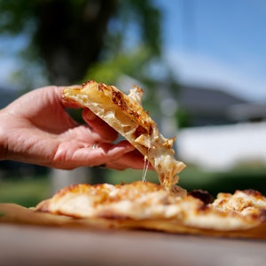 May include: A hand holding a slice of pizza with melted cheese and a crispy crust. The pizza is on a wooden board.