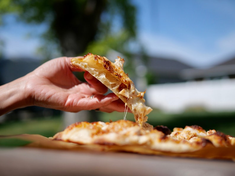 May include: A slice of pizza with melted cheese being held up, with the rest of the pizza in the background. The crust is golden brown and the cheese is stringy. The pizza is on a piece of brown parchment paper.
