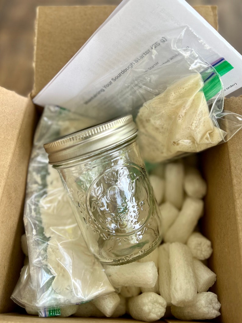 May include: A sourdough starter kit in a cardboard box. The kit includes a clear glass jar with a silver lid, packets of starter, and packing peanuts. A sheet of paper is visible with text.