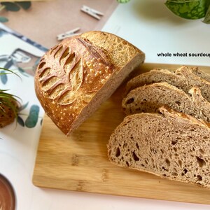 May include: A loaf of whole wheat sourdough bread, partially sliced, with a decorative leaf pattern on top. Several slices are arranged on a wooden cutting board. The text "whole wheat sourdough bread" is visible.