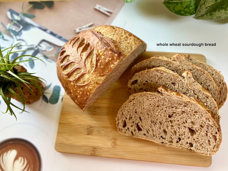 May include: A loaf of whole wheat sourdough bread, partially sliced, with a decorative leaf pattern on top. Several slices are arranged on a wooden cutting board. The text "whole wheat sourdough bread" is visible.