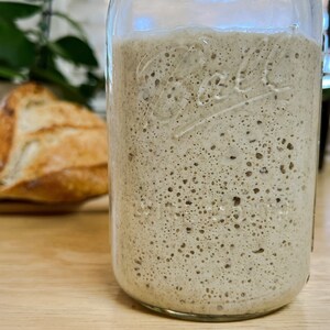 May include: A clear glass jar filled with bubbly, light brown sourdough starter. A loaf of crusty bread sits out of focus in the background on a wooden surface. The jar has embossed text.