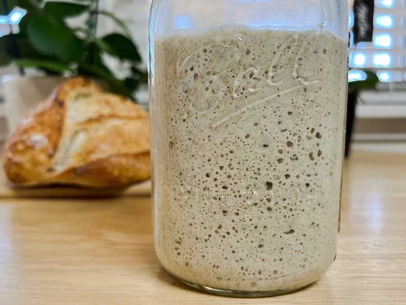 May include: A clear glass jar filled with bubbly, light brown sourdough starter. A loaf of crusty bread sits out of focus in the background on a wooden surface. The jar has embossed text.