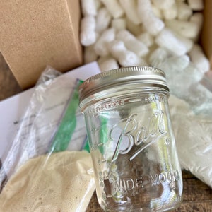May include: A clear glass jar with a silver lid and the text "Ball Wide Mouth" printed on the side. The jar is sitting on a brown wooden surface with packing peanuts in the background.