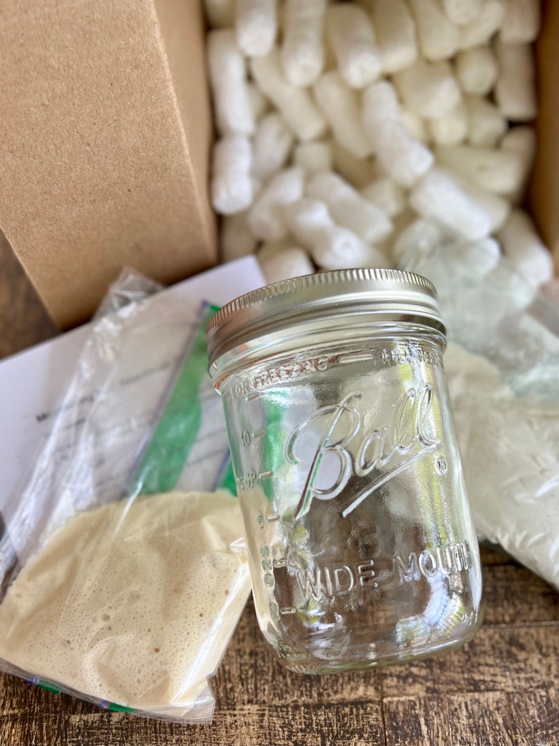May include: A clear glass jar with a silver lid and the text "Ball Wide Mouth" printed on the side. The jar is sitting on a brown wooden surface with packing peanuts in the background.