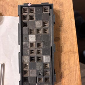 May include: A rectangular, black, open-top container with a grid of small, square compartments. The compartments are filled with small, dark gray, square blocks. The container is resting on a wooden surface.