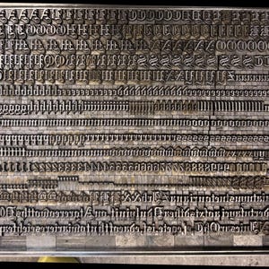 May include: A collection of metal letterpress type blocks arranged in rows on a flat surface. The grid features various gothic-style characters, including uppercase and lowercase letters, punctuation marks, and decorative symbols in silver-toned metal.