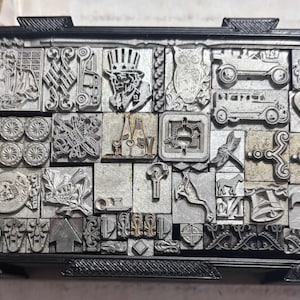 May include: A collection of vintage metal printing blocks in a black tray. The blocks feature various designs, including an Uncle Sam figure, vehicles, and decorative elements. The tray is rectangular with a raised edge.