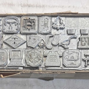 May include: A collection of vintage metal printing blocks, arranged in a wooden box. The blocks feature various designs, including floral patterns, stars, a car, a computer, and a telephone. The blocks are in a rectangular box.