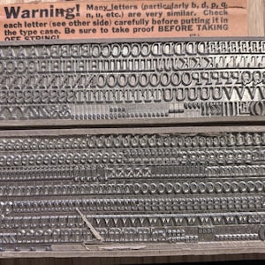 May include: Two rows of silver-colored metal letterpress type characters arranged in wooden blocks. A tan paper strip at the top reads: Warning! Many letters (particularly b, d, p, q, n, u, etc.) are very similar. Check each letter before putting it in the type case.