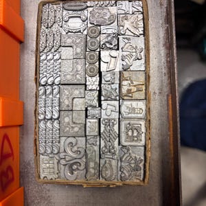 May include: A selection of vintage metal letterpress printing blocks, contained in a cardboard box. The blocks display a variety of decorative designs, including floral patterns, geometric shapes, and ornate borders. The blocks are silver and show signs of wear.