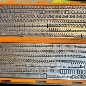May include: Two orange plastic trays filled with rows of metallic letterpress type characters. The silver-colored metal blocks are arranged in organized lines, featuring various uppercase and lowercase letters, punctuation marks, and symbols.