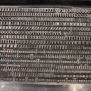 May include: A collection of metal letterpress type characters arranged in a rectangular grid. The set includes multiple rows of uppercase and lowercase letters in a serif font, all cast in dark grey metal and tightly packed together in a frame.