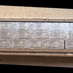 May include: A set of rectangular silver-colored metal blocks arranged in a horizontal row within a tan cardboard box. The blocks are stacked in two layers, featuring a metallic finish and a uniform, brick-like shape, neatly organized inside the container.