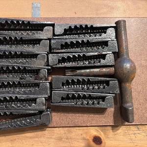 May include: A collection of vintage metal tools, including multiple wedge-shaped pieces with serrated edges and a handle. The tools are arranged on a brown surface, suggesting a workshop or craft setting.