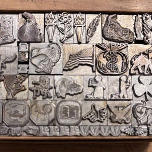 May include: A wooden tray filled with vintage metal printing blocks. The blocks feature various designs, including an American flag, an owl, a turkey, a dog, a shamrock, and other symbols. The blocks are arranged in a grid pattern.