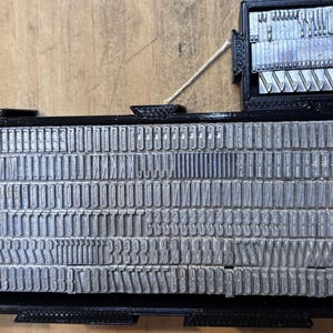 May include: A black 3D-printed storage tray containing rows of silver metal letterpress type characters. The tray features a smaller rectangular compartment attached to the top right, also filled with various metal type pieces.