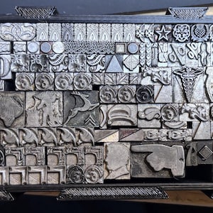 May include: A black rectangular tray filled with an assortment of metallic letterpress printing blocks. The collection includes various decorative motifs, geometric shapes, symbols, a pointing finger, a deer, and floral patterns in silver tones.