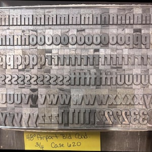 May include: A collection of metallic letterpress type blocks arranged in rows, featuring lowercase letters m through z and numbers. A yellow label at the bottom displays the handwritten text 48 pt Airport Bld Cond. 3/4 Case 620.