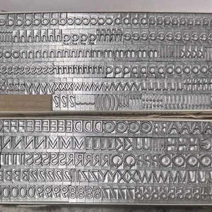 Puede incluir: Dos bandejas de tipos de impresión tipográfica de metal vintage. Las letras y los números están dispuestos en filas y columnas. El tipo es de metal de color plateado y se utiliza para la impresión.