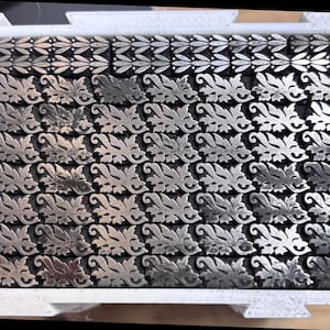 May include: A white rectangular tray containing a collection of small, metallic, silver-toned printing blocks. The blocks are arranged in rows and columns, featuring embossed leaf and scroll patterns, with a vertical column of leaf motifs on the right.