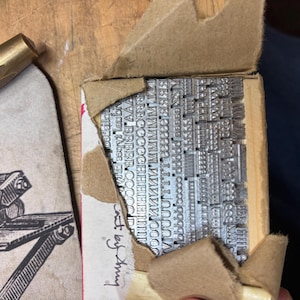 May include: A collection of silver-colored metal letterpress type characters arranged in a grid within a partially torn cardboard box. Next to the box is a light-colored card featuring a black and white illustration of a printing press.