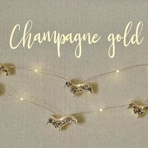 Puede incluir: Luces de cadena doradas champán con adornos de perro salchicha. Las luces están en un fino cable de cobre y tienen pequeñas luces LED blancas cálidas. El texto "Champagne gold" está escrito en cursiva.