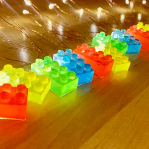 May include: A string of LED lights shaped like colourful Lego bricks. The lights are arranged in a rainbow pattern, with red, orange, yellow, green, blue, and purple bricks.