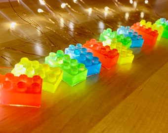 May include: A string of LED lights shaped like colourful Lego bricks. The lights are arranged in a rainbow pattern, with red, orange, yellow, green, blue, and purple bricks.