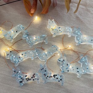 May include: A string of corgi-shaped fairy lights. The clear resin corgis are filled with silver and blue glitter. The lights are strung on a copper wire and are lit up with small, warm white lights.