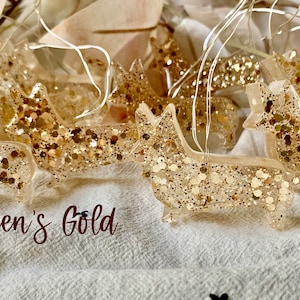 May include: A string of lights featuring gold glitter-filled resin ornaments shaped like dogs. The ornaments are strung together with clear wire and have small white lights. The text "Queen's Gold" is visible in the image.