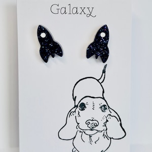 May include: A pair of black glitter rocket ship earrings on a white card with the word "Galaxy" printed on it.