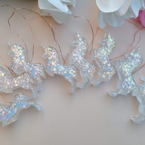 May include: A string of unicorn-shaped fairy lights. Each unicorn is clear with iridescent glitter, and they are connected by thin copper wire. The lights are arranged on a light-colored surface, with white and pink flowers in the background.