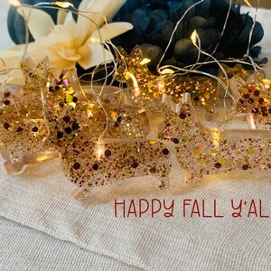 May include: A string of LED fairy lights featuring multiple translucent resin corgi-shaped ornaments filled with gold, copper, and dark red glitter. The lights are illuminated, and the text "HAPPY FALL Y'ALL" is printed in red at the bottom.