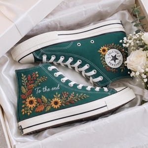 Può includere: Un paio di scarpe alte in tela verde scuro con lacci e suole in gomma bianche. Le scarpe sono ricamate con girasoli, foglie autunnali e il testo To the end. Sono presentate in una scatola regalo bianca con un nastro di raso bianco.