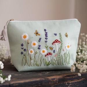 May include: A pale green cosmetic bag with a zip and tassel. The bag is embroidered with a garden scene featuring daisies, lavender, mushrooms, bees, and butterflies. The bag is on a wooden surface.