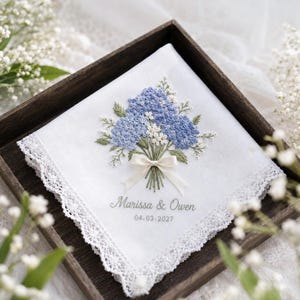 May include: A white square handkerchief with lace trim featuring an embroidered bouquet of blue and white flowers tied with a small cream ribbon. Below the flowers, the text reads "Marissa & Owen" followed by the date "04.03.2027" in grey script.