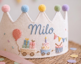 May include: A cream-colored fabric crown featuring the embroidered name Milo in blue script. The crown is decorated with colorful pom-poms on each point and embroidered motifs including a balloon, gift boxes, a birthday cake, and a drum.