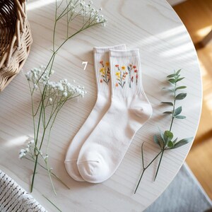 May include: A pair of off-white socks with embroidered floral designs in yellow, orange, and red. The socks are displayed on a light wooden surface, alongside sprigs of greenery and a woven basket.