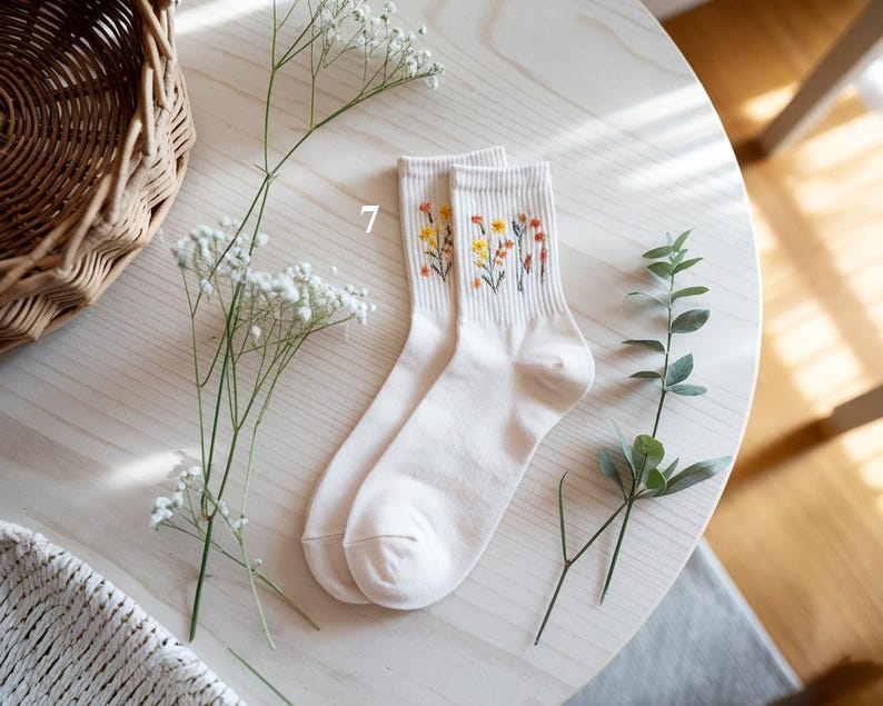 May include: A pair of off-white socks with embroidered floral designs in yellow, orange, and red. The socks are displayed on a light wooden surface, alongside sprigs of greenery and a woven basket.