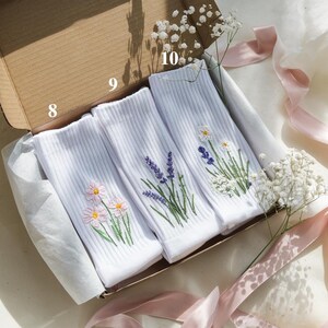 May include: Three pairs of white ribbed socks with embroidered floral designs, including daisies, lavender, and other wildflowers, are presented in an open cardboard box. The box is lined with white tissue paper and decorated with pink ribbons and baby's breath.