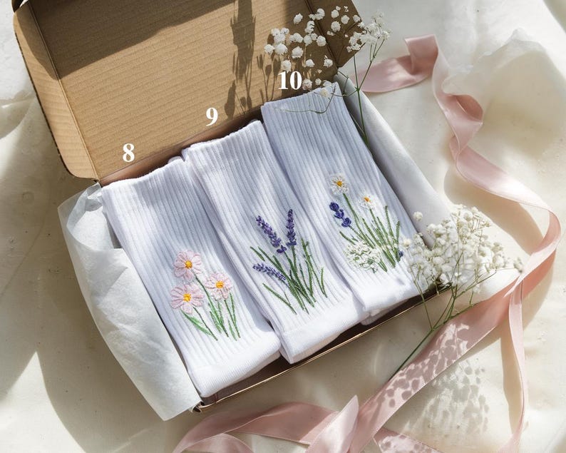 May include: Three pairs of white ribbed socks with embroidered floral designs, including daisies, lavender, and other wildflowers, are presented in an open cardboard box. The box is lined with white tissue paper and decorated with pink ribbons and baby's breath.