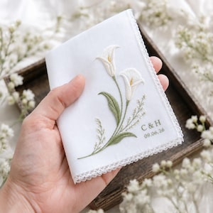Op de afbeelding: Een witte stoffen zakdoek met geborduurde witte calla lelies met groene stelen en bladeren. De hoek bevat de geborduurde tekst C & H en de datum 09.06.26 in groen garen. De randen zijn afgewerkt met een delicate witte kanten afwerking.