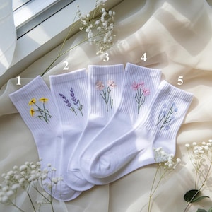 May include: Five pairs of white crew socks, each embroidered with a different floral design. The flowers include yellow, lavender, pink, and blue. The socks are arranged on a cream-colored fabric, with sprigs of baby's breath.