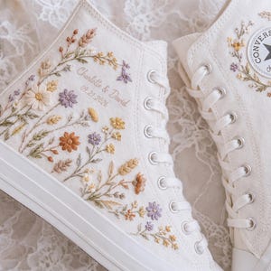 May include: A pair of off-white high-top canvas sneakers featuring intricate floral embroidery in shades of yellow, orange, and purple. The side displays the text Charlotte & David 09.21.2026, while the other shoe features the Converse All Star logo.